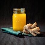 A mason jar filled with vibrant homemade orange ginger juice, placed on a dark fabric surface with a piece of fresh ginger root and a green cloth napkin.