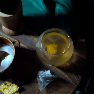 A cozy scene featuring a bag of tea, a steaming cup of brewed tea, a small bowl of sugar, and dehydrated pineapple slices on a wooden table.