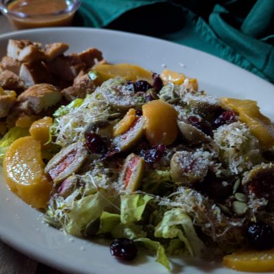 A colorful peach salad, featuring grilled chicken, juicy peach slices, green olives, parmesan cheese, pistachios, pumpkin seeds, dried cranberries, and a drizzle of peach glaze dressing. The mix of fresh ingredients creates a vibrant contrast of colors and textures, making for a visually stunning and delicious meal.