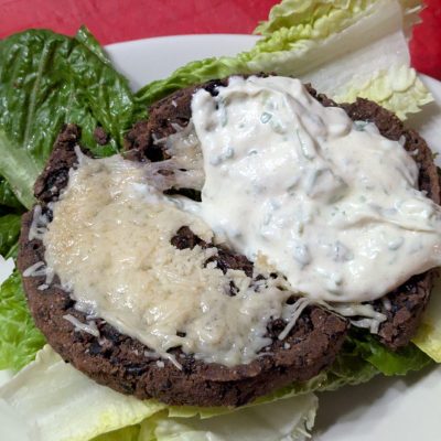 A hearty black bean burger served on a fresh bed of lettuce, topped with creamy cilantro dressing and a sprinkle of Parmesan cheese.