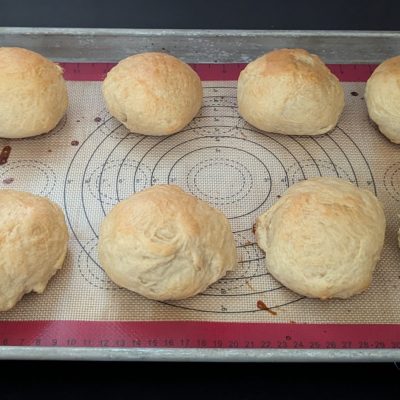 A tray of golden-brown, freshly baked dinner rolls resting on a sheet pan, their soft texture and fluffy interiors visible through slight cracks on the surface.
