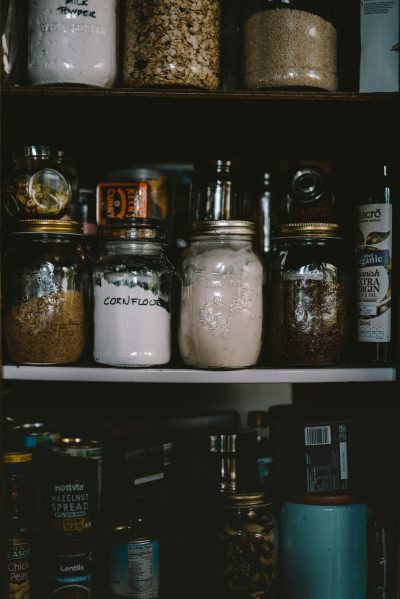 A well-stocked pantry with jars of dried beans, grains, pasta, canned goods, and spices neatly arranged on rustic wooden shelves.
